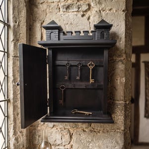 May include: A black wooden wall-mounted key cabinet shaped like a castle with a crenelated top. The cabinet door is open, revealing interior hooks holding three metal keys, with one additional key resting on the bottom shelf of the cabinet.