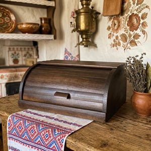 Puede incluir: Una panera de madera oscura con tapa curva y asa sobre una mesa de madera. Un paño estampado con diseños rojos y azules está sobre la mesa. Una pequeña maceta de terracota con hierbas secas y una cuchara de madera.