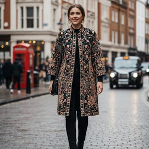 Pode incluir: Um casaco longo preto com bordados florais intrincados em tons de rosa, dourado e verde-azulado. O casaco apresenta uma gola mandarim e é usado sobre roupas pretas. O fundo mostra uma rua da cidade com edifícios.