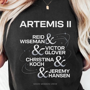 Puede incluir: Camiseta negra de cuello redondo con el texto "ARTEMIS II" y los nombres de la tripulación: Reid Wiseman, Victor Glover, Christina Koch y Jeremy Hansen. Las palabras "MOON MISSION CREW" están impresas en la parte inferior.