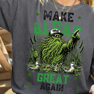 Könnte beinhalten: Ein anthrazitfarbenes T-Shirt mit einer Grafik einer grünen Sumpfkreatur, die eine Baseballkappe und eine Sonnenbrille trägt und nach oben zeigt. Der Text "MAKE ALGAE GREAT AGAIN" ist in Schwarz und Grün gedruckt. Zwei Enten schwimmen.