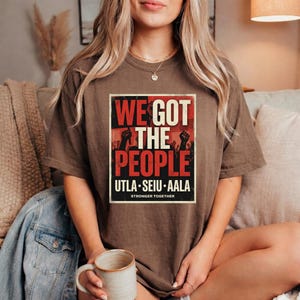 May include: A brown t-shirt with the text "WE GOT THE PEOPLE" in red and white, with black silhouettes of raised fists. The shirt also features the text "UTLA SEIU AALA" and "STRONGER TOGETHER" in a rectangular frame.