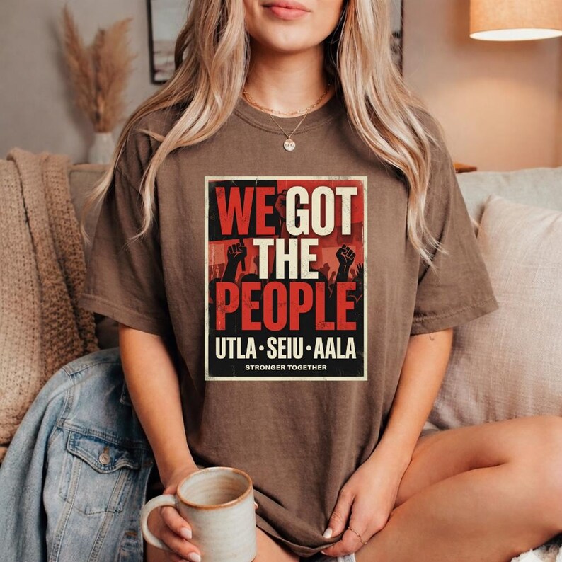 May include: A brown t-shirt with the text "WE GOT THE PEOPLE" in red and white, with black silhouettes of raised fists. The shirt also features the text "UTLA SEIU AALA" and "STRONGER TOGETHER" in a rectangular frame.