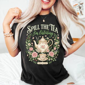 Könnte beinhalten: Schwarzes T-Shirt mit dem Text "Spill the Tea I'm listening" und einem floralen Design mit einer Teekanne, einer Teetasse und Rosen. Die Worte "Tea Lover" sind ebenfalls auf dem T-Shirt aufgedruckt.