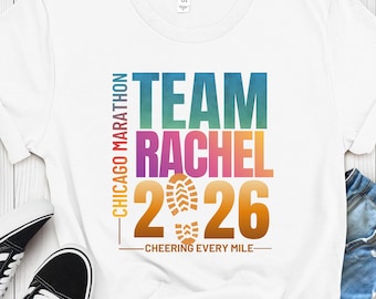 May include: A white short-sleeved t-shirt featuring a colorful graphic with the text "TEAM RACHEL 2026" in a gradient font, "CHICAGO MARATHON" vertically, and "CHEERING EVERY MILE" horizontally, with a central shoe print icon between the numbers.