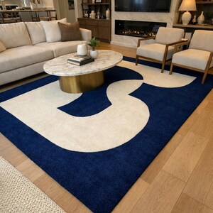 Könnte beinhalten: Ein rechteckiger Teppich mit einem auffälligen Design. Der Teppich hat einen marineblauen Rand und eine große, abstrakte cremefarbene Form in der Mitte. Der Teppich liegt auf einem Holzboden in einem Wohnzimmer.