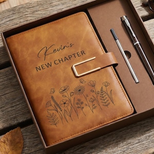 May include: A tan leather-like journal with a gold-tone metal clasp, featuring the text "Kevin's NEW CHAPTER" and a black floral line art design. The journal is housed in a brown gift box alongside a silver pen and a separate ink refill cartridge.