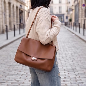 Peut inclure: Un sac à bandoulière en cuir marron clair doté d'un grand rabat rectangulaire, d'une fermeture à verrou tournant en métal argenté et d'une bandoulière réglable. Le sac présente un design structuré et lisse avec des finitions argentées.