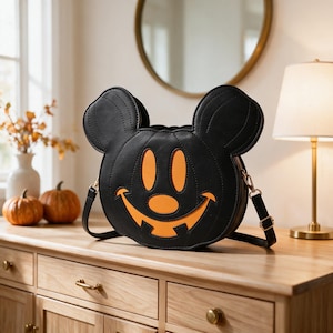 May include: A black circular crossbody bag shaped like a mouse head with large rounded ears. The front features a bright orange jack-o'-lantern face cutout with a wide smiling mouth, two oval eyes, and a small nose. It has a thin black shoulder strap.