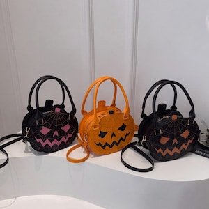 May include: Three round handbags shaped like jack-o'-lanterns, each featuring a top handle and a shoulder strap. The left bag is black with pink details, the center bag is orange with black details, and the right bag is black with orange details.