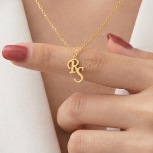 May include: A gold-coloured necklace featuring the initials "RS" in a delicate, cursive font. The pendant hangs from a fine chain. The necklace is displayed against a white background.