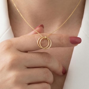 May include: A delicate gold necklace featuring two interlocking circles. The pendant hangs from a fine chain, creating a minimalist and elegant design. The necklace is shown against a white blazer, highlighting its subtle shine and simple beauty.