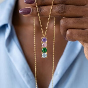 May include: A gold-tone necklace featuring a vertical bar pendant set with four round-cut gemstones in purple, pink, green, and light blue. The pendant hangs from a delicate gold-tone chain, creating a layered look with the thin metal links.