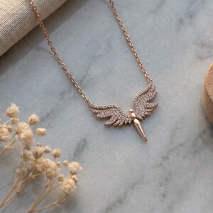 May include: Rose gold-toned necklace featuring a pendant shaped like angel wings. The wings are encrusted with small, sparkling stones. The necklace is displayed on a marble surface with a wooden tray and dried flowers in the background.