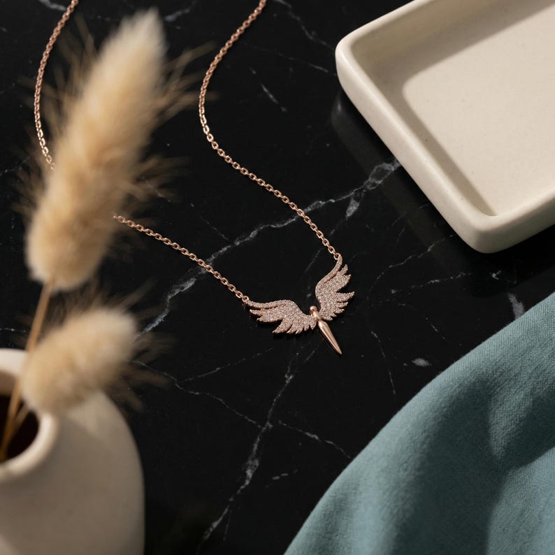 May include: A rose gold necklace featuring an angel pendant with crystal-like accents. The angel has outstretched wings and a pointed lower body. The necklace is displayed on a black marble surface with a white ceramic dish and a sprig of dried flowers.