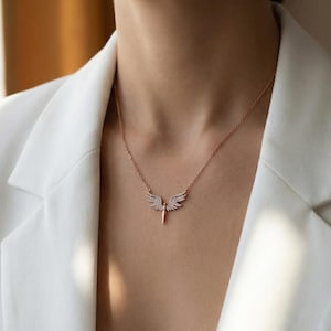 May include: A rose gold necklace featuring a pendant with a wing design. The wings are encrusted with small, sparkling crystals. The necklace is worn against a white blazer.