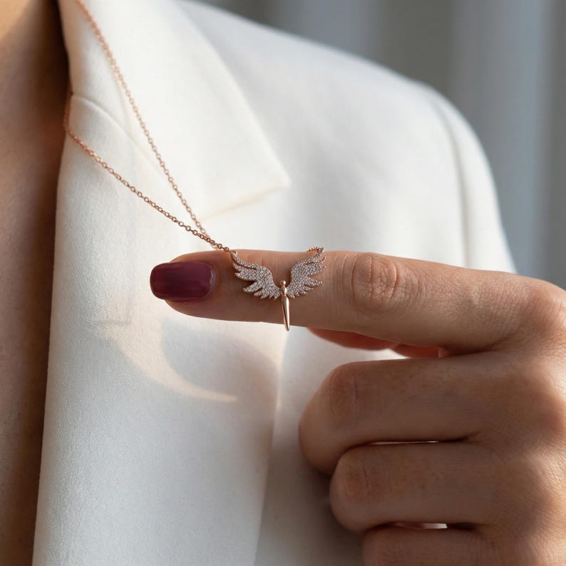 May include: Rose gold necklace featuring a pendant with angel wings and a central figure. The wings are encrusted with small, sparkling stones. The necklace is displayed against a white background.