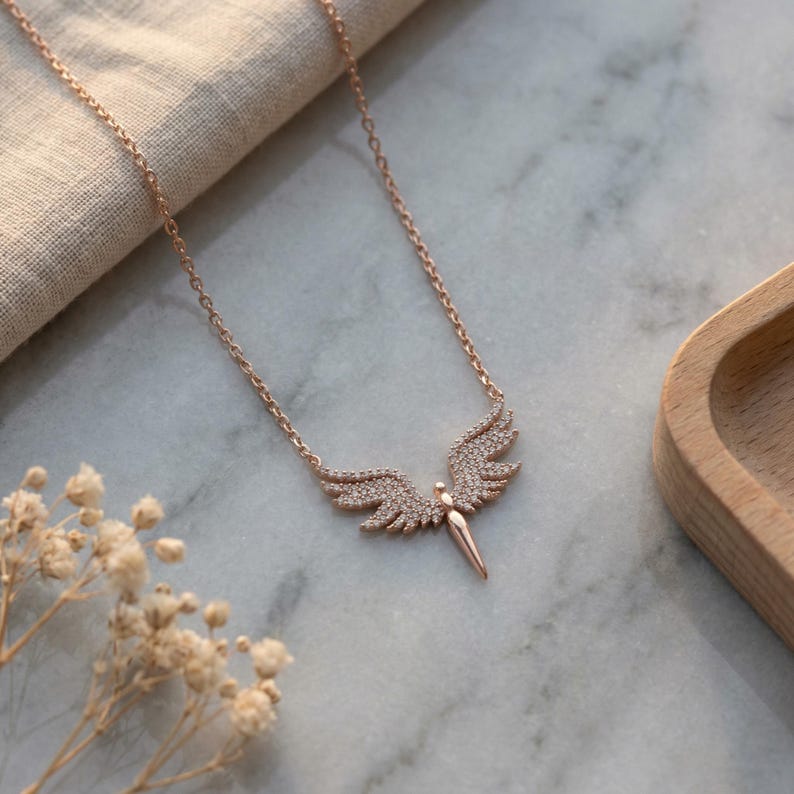 May include: Rose gold-toned necklace featuring a pendant shaped like angel wings. The wings are encrusted with small, sparkling stones. The necklace is displayed on a marble surface with a wooden tray and dried flowers in the background.