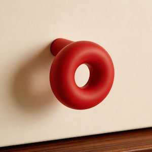 May include: A red, ring-shaped drawer pull is attached to a cream-coloured cabinet. The pull is a vibrant red and has a circular design with a hole in the centre. The cabinet is set above a dark wood surface.