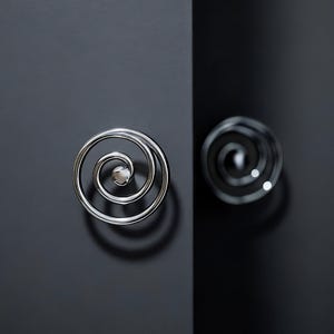 Peut inclure: Deux poignées de placard argentées en forme de spirale sur un fond gris foncé. Les poignées ont une finition polie et un petit centre rond. Les poignées sont montées sur une porte de placard gris foncé.