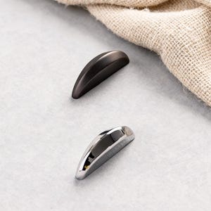 Puede incluir: Dos tiradores de armario curvos, uno en color bronce oscuro mate y otro cromado brillante. Los tiradores se colocan sobre un fondo gris neutro con una tela beige en la esquina superior derecha.