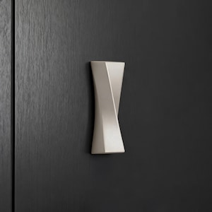 Può includere: Una maniglia per armadio attorcigliata beige chiaro con un design geometrico. L'hardware è montato su un'anta di un armadio in legno grigio scuro. La maniglia ha un'estetica moderna e minimalista ed è probabilmente in metallo.