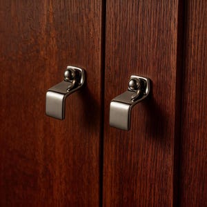 Może przedstawiać: Zbliżenie dwóch drzwi szafki z okuciami ze szczotkowanego niklu. Okucia mają zakrzywioną konstrukcję uchwytu z małym detalem w kształcie kuli na górze. Drzwi wykonane są z ciemnego, bejcowanego drewna.