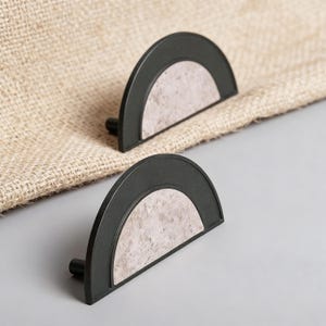 Op de afbeelding: Twee halfronde kastgrepen. Elke greep heeft een donkergrijs, gebogen frame met een lichtgrijs, getextureerd midden. De grepen zijn geplaatst tegen een neutraal getinte, getextureerde achtergrond en een grijs oppervlak.