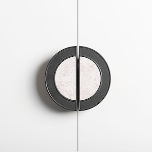 Op de afbeelding: Twee zwarte kastgrepen met een cirkelvormig ontwerp. Elke greep heeft een licht, gespikkeld midden en een donkere buitenring. Ze zijn gemonteerd op een wit oppervlak, met een dunne zwarte lijn ertussen.