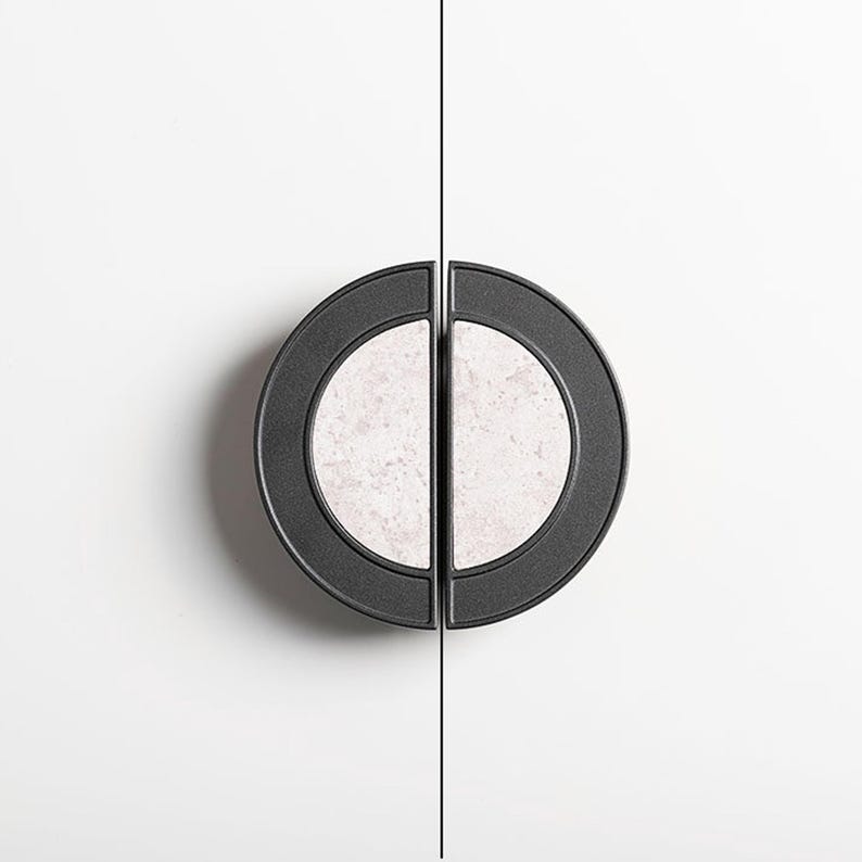 Op de afbeelding: Twee zwarte kastgrepen met een cirkelvormig ontwerp. Elke greep heeft een licht, gespikkeld midden en een donkere buitenring. Ze zijn gemonteerd op een wit oppervlak, met een dunne zwarte lijn ertussen.