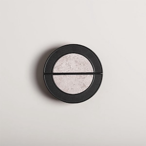 Op de afbeelding: Een rond, zwart object met een lichtgrijs, steenachtig midden. Het midden is verdeeld in twee halve cirkels. Het object staat tegen een witte achtergrond, wat een minimalistische esthetiek cre&euml;ert.