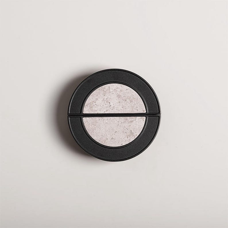Op de afbeelding: Een rond, zwart object met een lichtgrijs, steenachtig midden. Het midden is verdeeld in twee halve cirkels. Het object staat tegen een witte achtergrond, wat een minimalistische esthetiek cre&euml;ert.