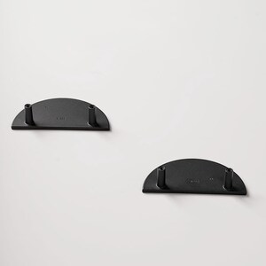 Op de afbeelding: Twee zwarte, halfronde metalen plankbeugels met elk twee cilindrische palen. De beugels zijn op een witte achtergrond geplaatst. De beugels zijn waarschijnlijk bedoeld om planken of andere decoratieve items te ondersteunen.