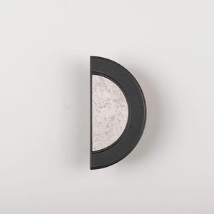 Op de afbeelding: Een zwarte, halfronde kastgreep met een lichtgrijs, gespikkeld midden. De hardware staat tegen een effen witte achtergrond. Dit moderne ontwerp is geschikt voor laden en kasten.