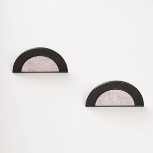 Op de afbeelding: Twee halfronde wandplanken. Elke plank heeft een zwart frame en een lichtgrijs, getextureerd interieur. De planken zijn op een witte muur gemonteerd, wat een minimalistische en moderne esthetiek cre&euml;ert.