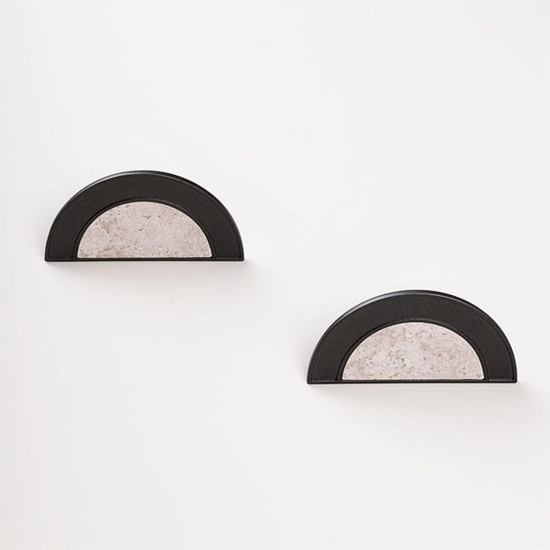 Op de afbeelding: Twee halfronde wandplanken. Elke plank heeft een zwart frame en een lichtgrijs, getextureerd interieur. De planken zijn op een witte muur gemonteerd, wat een minimalistische en moderne esthetiek cre&euml;ert.