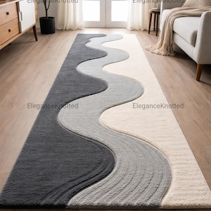 Puede incluir: Una alfombra larga y rectangular con un diseño ondulado en tonos grises y crema. La alfombra presenta una sección gris oscuro, una sección gris medio y una sección color crema, creando un patrón fluido y texturizado. La alfombra está colocada sobre un suelo de madera.