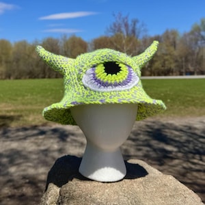 May include: A lime green crochet bucket hat with a large, central eye design. The eye features a black pupil, a yellow iris, and purple and white accents. The hat has two small, upward-curving horns.