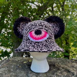 May include: A crochet bucket hat in a black and white variegated yarn featuring two large, fuzzy black pom-poms on the sides and a prominent, centred eye appliqué with a bright pink iris, a black pupil, and a white border on the front panel.