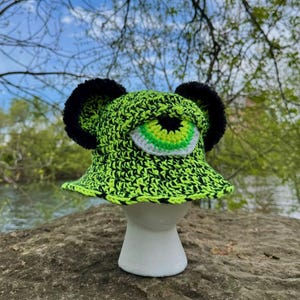 May include: A neon green and black crochet bucket hat with a large, cartoonish eye and black pom-poms. The eye features white, green, and black details. The hat is displayed on a white mannequin head against a natural outdoor backdrop.