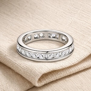 Puede incluir: Un anillo de eternidad plateado con una fila continua de piedras preciosas redondas y claras. El anillo está colocado sobre una tela beige, mostrando su metal pulido y piedras brillantes. Una joya clásica.
