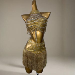 以下が含まれることがあります： 金色のダンスコスチューム。複雑なビーズとフリンジが特徴です。ホルタースタイルのトップスとスカートには、ラインストーンとチェーンが装飾されています。スカートには長いフリンジが付いています。