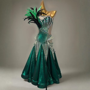 May include: Emerald green ballroom dance dress with a full skirt and embellished bodice. The dress features a one-shoulder design with a feather accent and is adorned with rhinestones and pearl detailing. The dress is displayed on a mannequin.