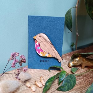 Peut inclure: Une broche en bois en forme d'oiseau avec un motif floral sur une carte bleue. L'oiseau a un corps brun clair, un bec jaune et un motif floral coloré sur son aile. Le mot "BROCHE" est imprimé en blanc en dessous.