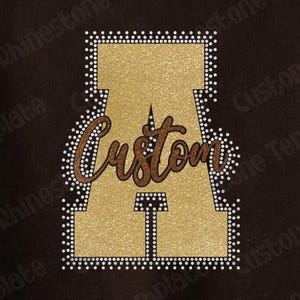 May include: A large gold glitter letter A outlined with small white rhinestones. The word Custom is written across the center of the letter in a brown glitter script font. The entire design is set against a dark brown background.