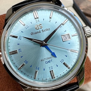 May include: A Grand Seiko automatic GMT watch with a light blue sunray dial, silver-tone indices, and a bright blue GMT hand. The watch features a date window at the 3 o'clock position, a black leather strap, and a polished stainless steel case.