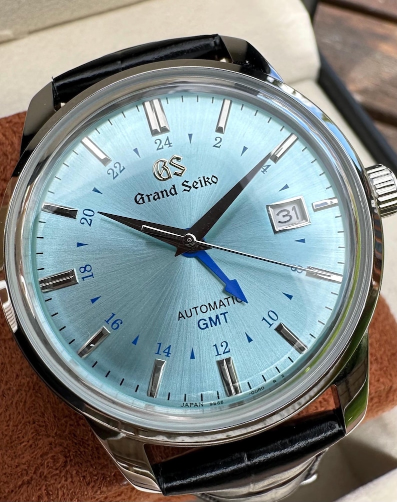 May include: A Grand Seiko automatic GMT watch with a light blue sunray dial, silver-tone indices, and a bright blue GMT hand. The watch features a date window at the 3 o'clock position, a black leather strap, and a polished stainless steel case.