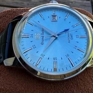 May include: A Grand Seiko automatic GMT watch with a light blue sunray dial, silver-tone indices, and a date window at the three o'clock position. The timepiece features a polished stainless steel case and a black leather strap with visible stitching.