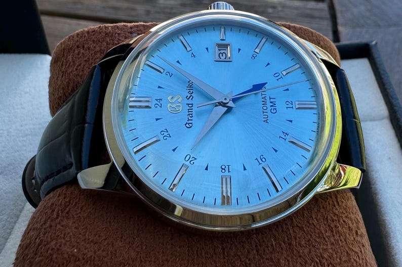 May include: A Grand Seiko automatic GMT watch with a light blue sunray dial, silver-tone indices, and a date window at the three o'clock position. The timepiece features a polished stainless steel case and a black leather strap with visible stitching.