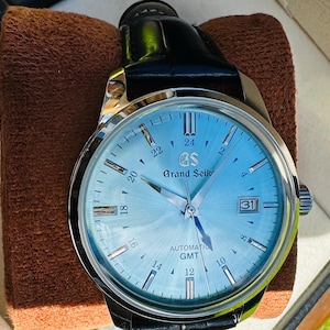 May include: A Grand Seiko automatic GMT watch with a light blue sunray dial, silver-tone indices, and a date window at the three o'clock position. The timepiece features a polished stainless steel case and a black leather strap with a buckle.